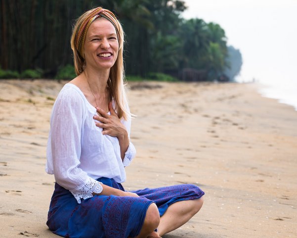 Eine blonde Frau sitzt lachend an einem Strand