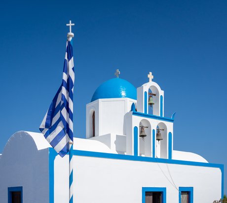 Eine weiße Kirche mit blauem Rand und einem runden Dach mit griechischer Flagge davor