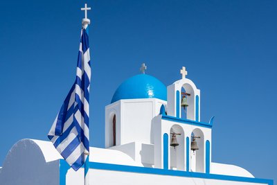 Eine weiße Kirche mit blauem Rand und einem runden Dach mit griechischer Flagge davor