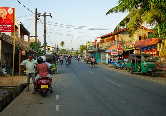 Eine Straße mit Palmen und verschiedenen Häusern und roller-fahrende Menschen