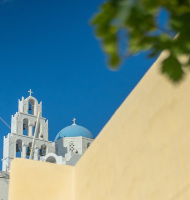 Hinter einer gelben Mauer befindet sich eine weiße Kirche mit sechs Glocken