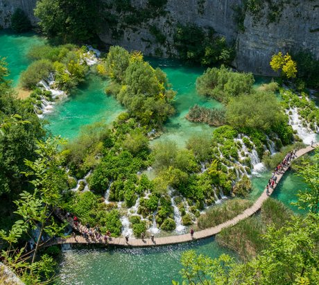 Ein See mit vielen kleinen Wasserfällen und Bäumen durch den ein Steg führt