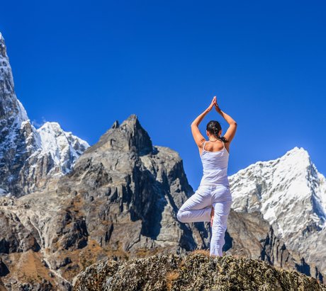 Eine junge Frau in weiß gekleidet steht in Baum-Pose auf das Gebirge des Himalaya blickend