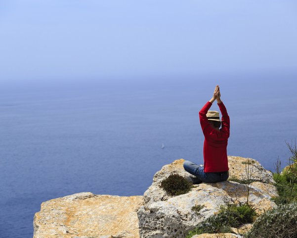 Ein Mensch sitzt auf einem Felsen über dem Meer die Hände im Anjali Mudra über dem Kopf