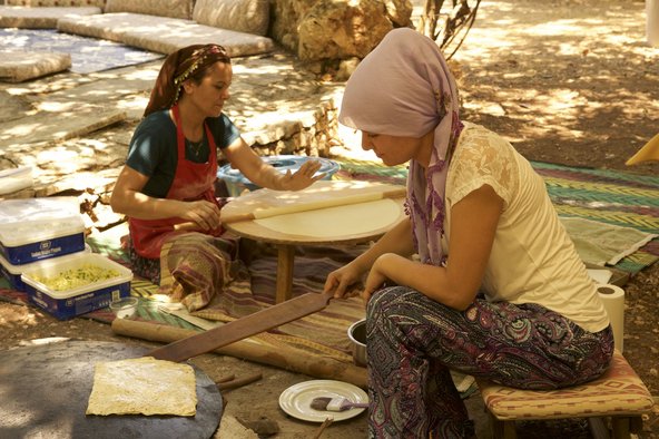 Zwei Frauen sitzen auf dem Boden vor Tisch und Pfanne und backen eine Spezialität aus Teig