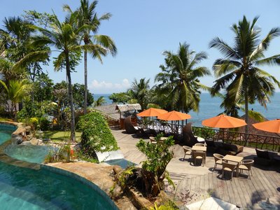 Vom Pool-Deck des Puri Dajuma blicken Sie über den leise plätschernden Pool durch grüne Palmen zum Meer