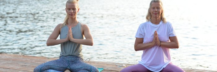 Zwei blonde Frauen sitzen in Meditationshaltung auf einem Steg am Meer
