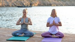 Zwei blonde Frauen sitzen in Meditationshaltung auf einem Steg am Meer
