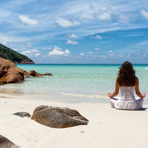 Eine Frau sitzt im Schneidersitz an einem Strand direkt am Meer