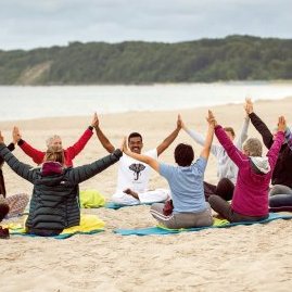 Mehrere Menschen sitzen in einem Kreis am Strand und heben die Hände über den Kopf