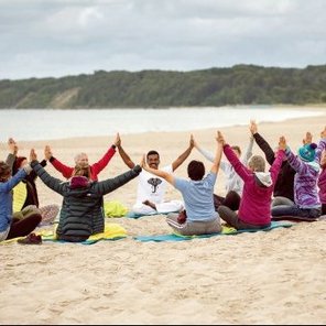 Mehrere Menschen sitzen in einem Kreis am Strand und heben die Hände über den Kopf